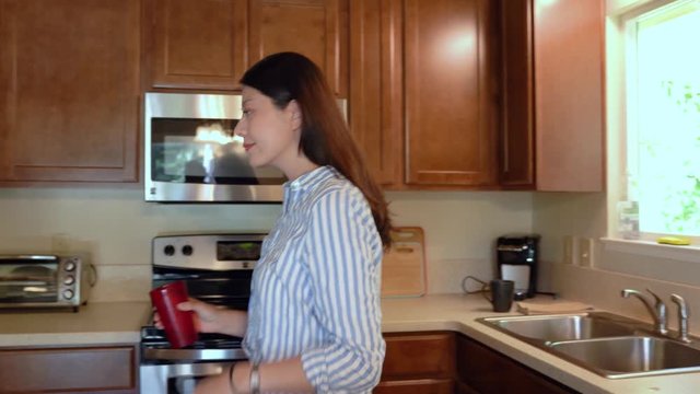young beautiful thirsty asian woman relax at home walking fast in wooden kitchen opening cupboard take out red cup and pressing into modern refrigerator water dispenser filling for refreshing drink.