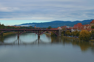 Fototapeta premium Autumn view of Drava River in Maribor. Ancient metal arch bridge, named Old Bridge, connects two parts of the city. Famous touristic place and romantic travel destination. Maribor, Slovenia