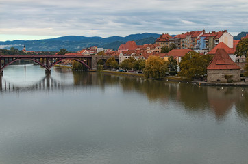 Fototapeta premium Tranquil autumn morning in Maribor. Beautiful Old Bridge reflected in water of the Drava River. Ancient building with red tile roofs at the background. Travel and tourism concept. Maribor, Slovenia
