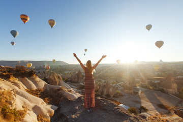 Young attractive girl in a hat stands on the mountain with flying air balloons on the...