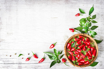 Red fresh chili peppers in wooden bowl on white old table. Spicy or hot small pepper with green leaves top view and copy space.