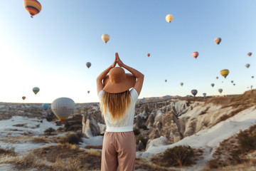 Beautiful young attractive girl in a hat stands on the mountain with flying air balloons on the background. Girl in the sunrise. View from the back. Famous tourist Turkish region Cappadocia.