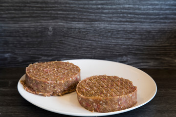 Single white plate with freshly pressed homemade burger patties. Sitting on dark wooden bench. Pattys are stuffed with bacon and cheese using burger stuffer. Meat is ground minced beef and pork mixed.