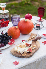 Outdoor standing decorated table with cheese platter on wooden board, pumpkins, grapes, figs, burning candles, colored glasses, bottle of wine and red autumn maple leaves.