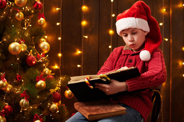 Teen boy reading book, sitting indoor near decorated xmas tree with lights, dressed as Santa helper - Merry Christmas and Happy Holidays!