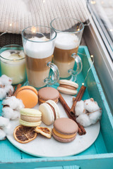 Turquoise breakfast tray with two cups of latte macchiato, macarons, cotton flowers, cinnamon rolls, dried orange slices, burning candle and string lights. Knitted sweaters on the background.