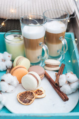 Turquoise breakfast tray with two cups of latte macchiato, macarons, cotton flowers, cinnamon rolls, dried orange slices, burning candle and string lights. Knitted sweaters on the background.