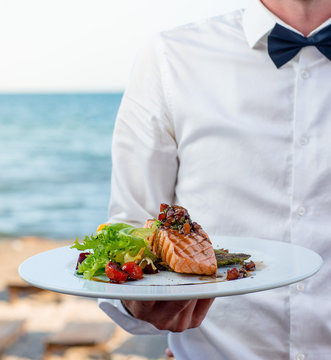 waiter holds a plate of grilled smoked salmon with lettuce, tomato, pepper
