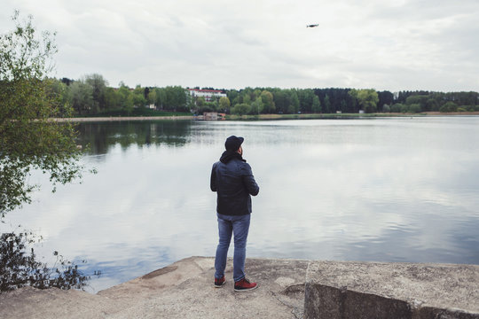 a man in a leather jacket controls a drone in the background of the lake
