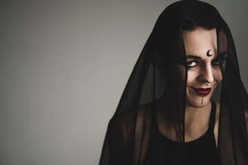 Redhead woman with a black veil on her head in a dark studio. A girl with an earring in her nose smiles slyly. Witch Makeup. Halloween costume.