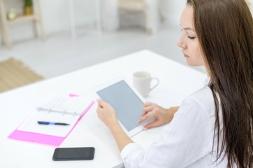 A young woman in a white shirt and jeans sits at a table with a tablet in her hands. A female office manager performs work on a modern device. The view from behind.