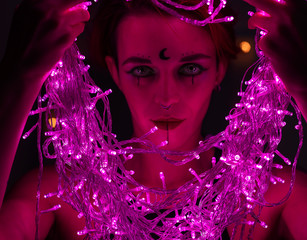 Woman with witch makeup and an earring in her nose. The girl holds a garland of flickering pink lights near her face. Light bulbs illuminate the witch's face in the dark.