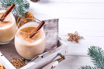 Winter spicy hot drink eggnog in a glasses with cinamon in tray, dry orange,fir branches and pine...