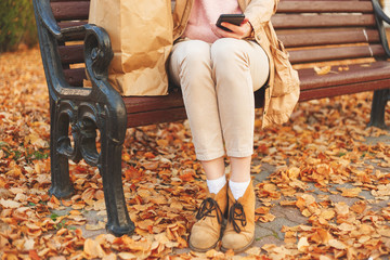 Young lady sitting on bench with paper bag and phone in autumn park