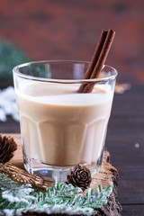 Eggnog Christmas milk cocktail with cinnamon, served in two glasses on a wood cutting board with fir branch and pine cone on dark wooden background
