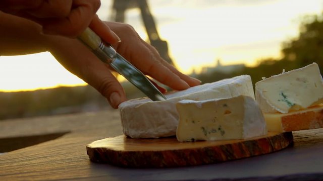 Hand slices soft brie cheese into sections on a wooden board on the background of the Eiffel Tower, Paris, France