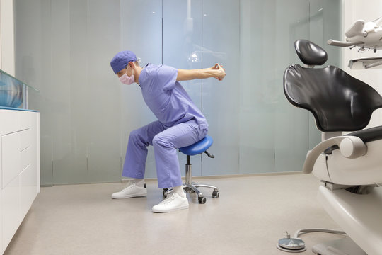 Dentist in uniform and mask, stretching arms in his office - healthy lifestyle at work