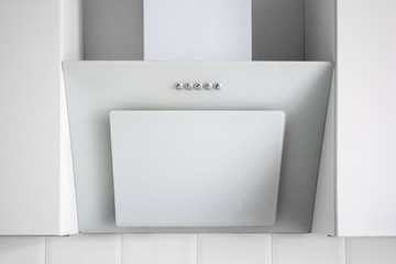 White tilted two-layer cooker hood with five silver buttons, located between two hanging cabinets. Simple light modern kitchen design