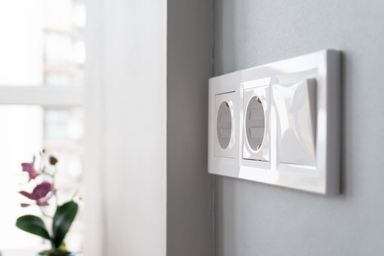 A closeup view of a group of white european electrical outlets and a switch located on a gray wall in a light modern kitchen by the window. Selective focus. Blurred background