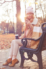 Obraz na płótnie Canvas Redhead girl in warm clothes sitting with smartphone on wooden bench in autumn park.