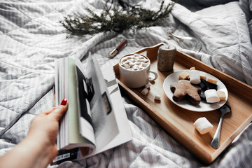 tray with a Cup of coffee with marshmallows, a saucer of cookies and marshmallows on a light blanket, a candle, a fir branch, a magazine in a woman's hand