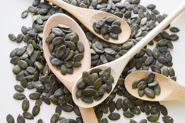 Uncooked organic pumpkin seeds in wooden spoons on white background, top view