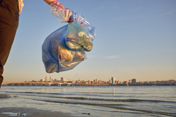 Young volunteer in purple gloves is walking with garbage bag along a dirty beach of the river and cleaning up trash. People and ecology. Close-up.