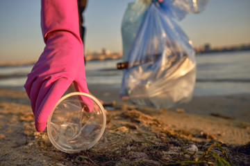 Young volunteer in purple gloves is walking with garbage bag along a dirty beach of the river and cleaning up trash. People and ecology. Close-up.