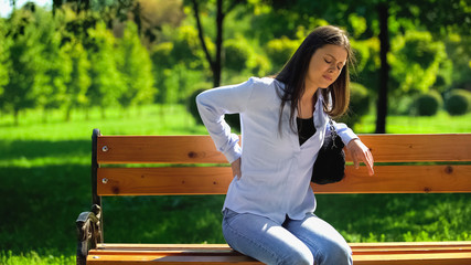Woman with sharp back pain sitting on bench in park to rest, sedentary lifestyle