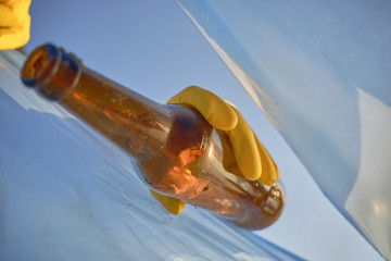 Hand of a conscious person in a yellow rubber glove puts a used brown glass bottle in a blue trash bag. Blue sky. Riverside pollution. Close-up shot.