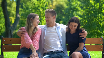 Handsome man resting on bench in park hugging two attractive females, ladies man