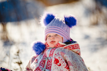 portrait A small child in a winter hat with blue pompons looks away. laughs. sunny weather. snow falls. age 2 years