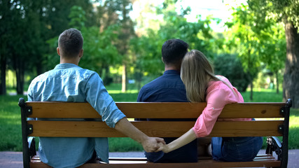 Couple hugging on bench, woman secretly holding hand of other man, betrayal