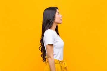 young pretty latin woman on profile view looking to copy space ahead, thinking, imagining or daydreaming against orange wall