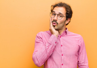 young handsome man feeling shocked and astonished holding face to hand in disbelief with mouth wide...