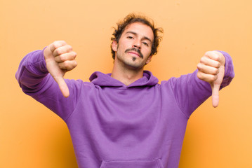 young handsome man looking sad, disappointed or angry, showing thumbs down in disagreement, feeling...