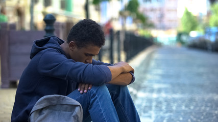 Upset multi-ethnic boy lonely sitting on sidewalk, family conflict, rebellion