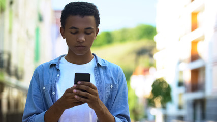 Afro-American male teenager chatting smartphone outdoors, online communication