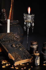 Books with spells, old pot, jars of potion are on a wooden dark table. Petals of dried roses are scattered nearby. Black background. Close-up shot.