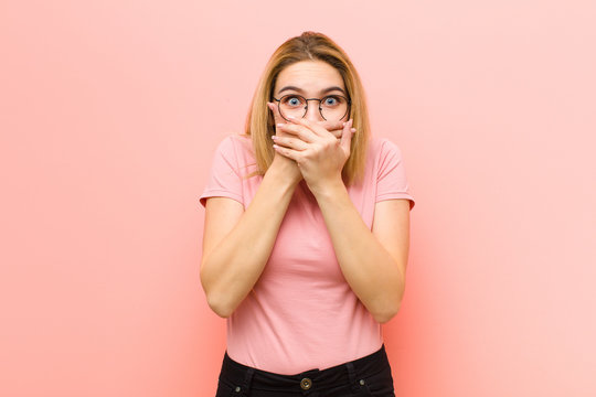 young pretty blonde woman covering mouth with hands with a shocked, surprised expression, keeping a secret or saying oops against pink flat wall
