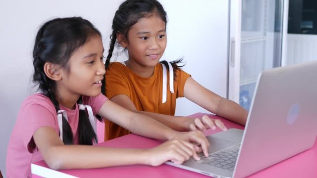 The older sister, who is wearing a pink T-shirt, is using internet for searching knowledge on laptop with young sister who is wearing an orange T-shirt. Both of them braid and cute white ribbons.