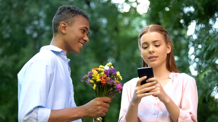 Indifferent teen girl ignoring boyfriend presenting flowers, chatting smartphone