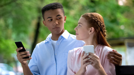 Mixed-race teen couple looking at each other chatting on smartphone in park