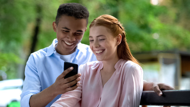 Smiling couple of teenagers watching photos on smartphone sitting on bench