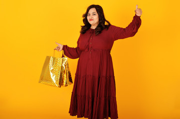 Attractive south asian woman in deep red gown dress posed at studio on yellow background with golden shopping bags.