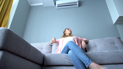 Young woman sitting on couch under air conditioner, cooling down on hot summer
