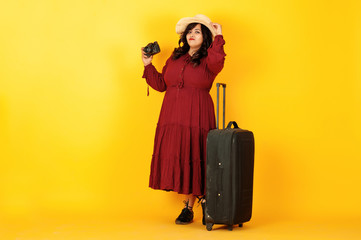 Attractive south asian traveler woman in deep red gown dress, hat posed at studio on yellow background with suitcase amd old vintage photo camera.