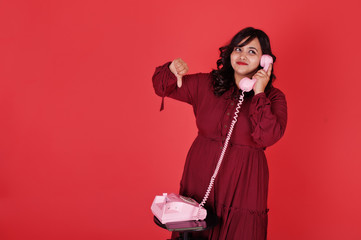 Attractive south asian woman in deep red gown dress posed at studio on pink background and use old vintage telephone and show thumb down.