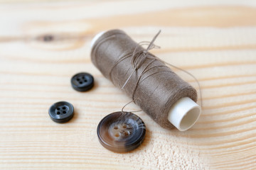 A bobbin of brown threads with a needle and three buttons lie on a wooden background