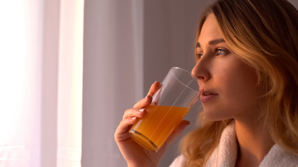 Girl in robe drinks orange juice looking in window, vitamins for skin, face care